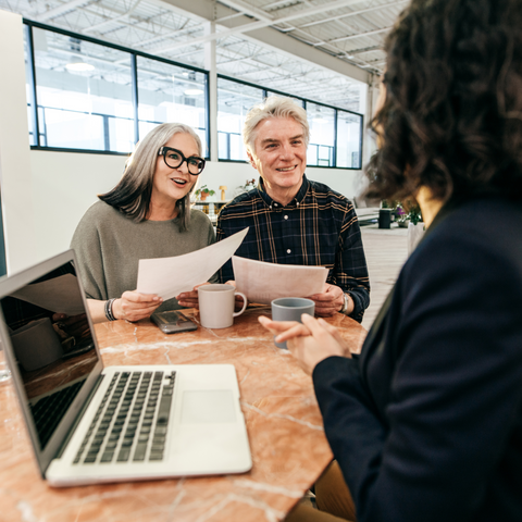 "A professional woman sitting at a table with an elderly couple, discussing documents. The woman is holding a piece of paper, and the couple is looking at the papers with cups of coffee in front of them. A laptop is on the table."