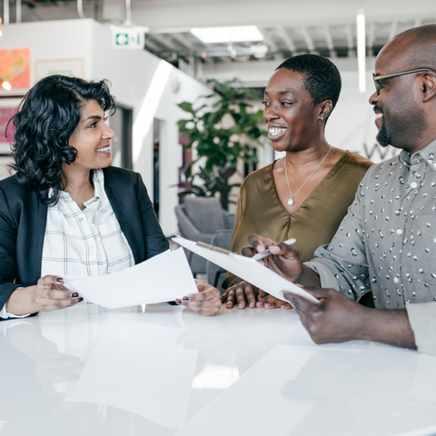 Three diverse professionals, two women and one man, engaged in a discussion while looking at documents in a modern office setting. They are smiling and appear to be collaborating on a project.