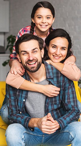 "A happy family of three, with a father, mother, and daughter, all smiling and hugging together. The father is sitting with the daughter on his lap, and the mother is behind them, with her arms around both of them."