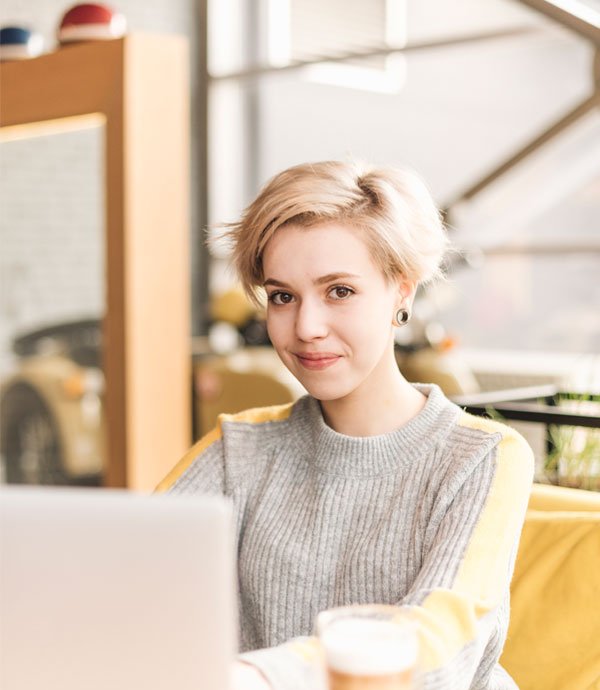 A young woman with short blonde hair, wearing a gray sweater, sitting at a table with a laptop in front of her and a cup of coffee. She is smiling and appears relaxed, in a cozy indoor setting.