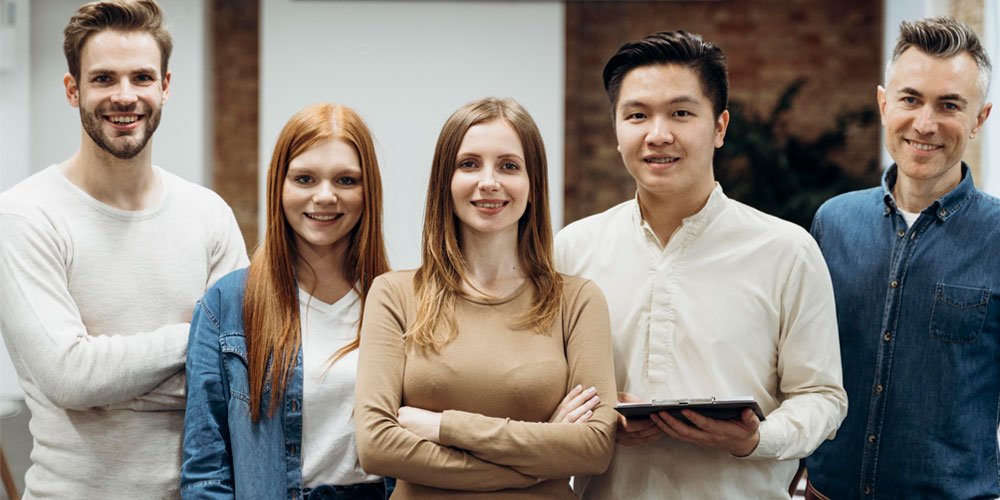 A diverse group of four people smiling and standing together, in a modern office setting. They appear friendly and professional, with two men and two women dressed in casual business attire.